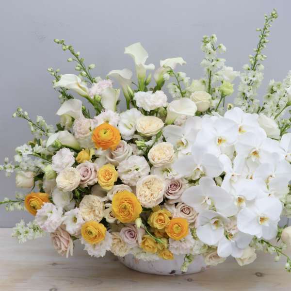 Large low arrangement of white orchids, calla lilies, and yellow and cream roses in a white bowl.