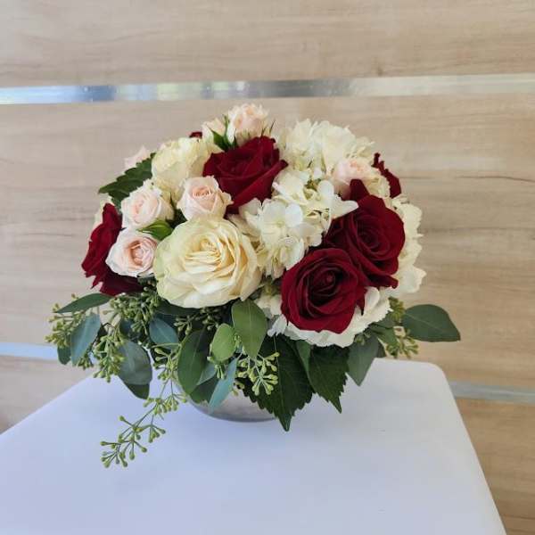 Low round arrangement of red and cream roses, blush spray roses, and white hydrangeas in a small bowl vase.