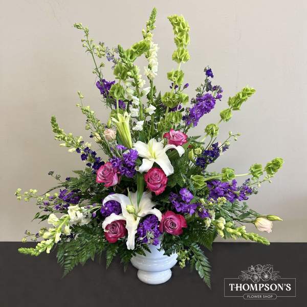 Tall fan-shaped arrangement with white lilies, pink roses, purple blooms, and green bells of Ireland in a white urn.