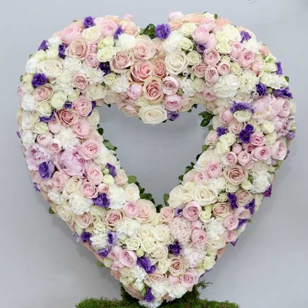 Heart-shaped standing arrangement of pink and white roses with purple accents on a moss-covered base.