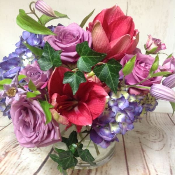 Low glass vase of purple roses, blue hydrangeas, and red amaryllis blooms in a compact arrangement