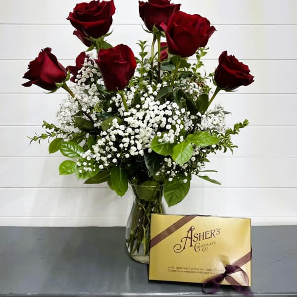 Gift set with red roses and white filler flowers in a vase beside a gold box of chocolates.