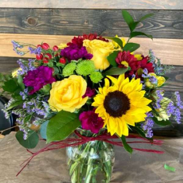 Mixed bouquet with yellow sunflower, yellow roses, and purple flowers in a clear glass jar