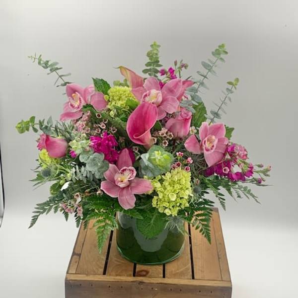 Pink calla lilies, orchids, and green hydrangeas in a clear glass vase on a wooden crate