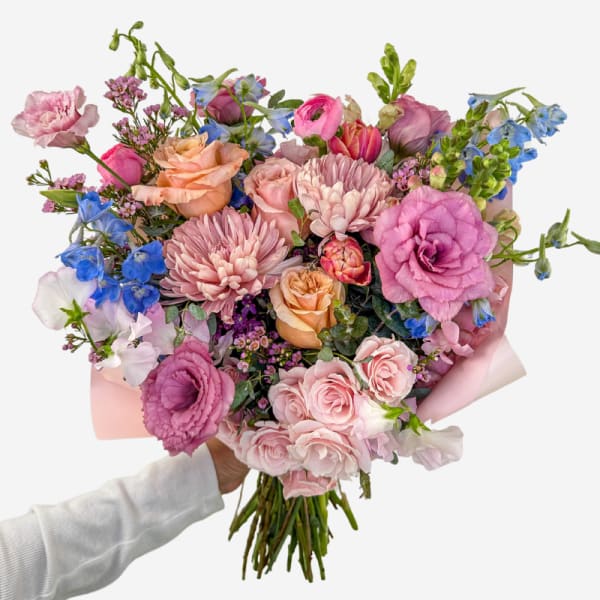 Handheld bouquet of pink, peach, and blue flowers wrapped in pale paper