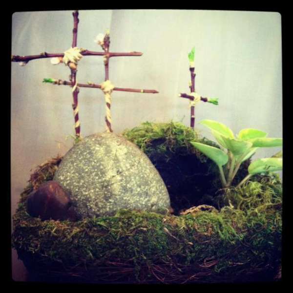 Moss-covered planter with small twig crosses and a green plant