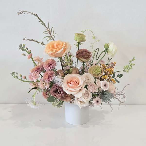 Asymmetrical arrangement of peach and blush flowers in a white cylinder vase