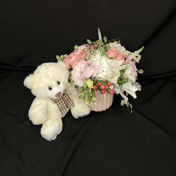 White teddy bear beside a pink floral arrangement in a ribbed vase