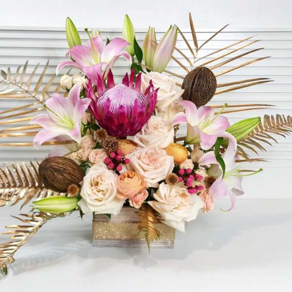 Modern arrangement with pink lilies, roses, a central protea and gold-painted leaves in a square glass vase