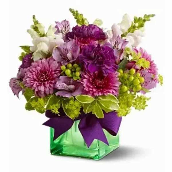 Compact arrangement of purple and pink flowers in a green cube vase with a purple bow