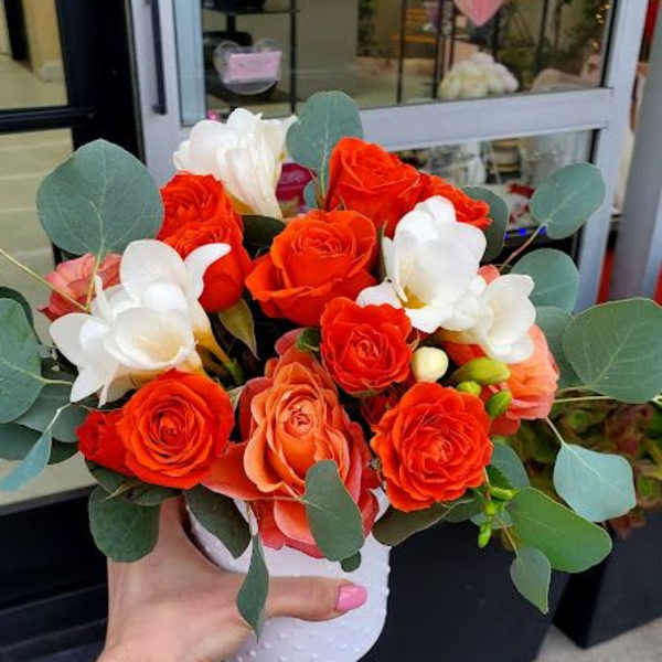 Compact orange and white rose arrangement in a white pot held outside a flower shop