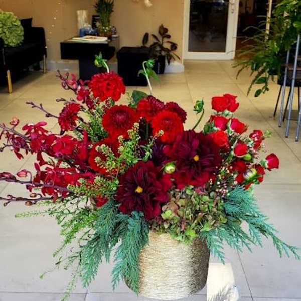 Low, lush arrangement of red roses and deep red flowers with evergreens in a gold container on a clear stand