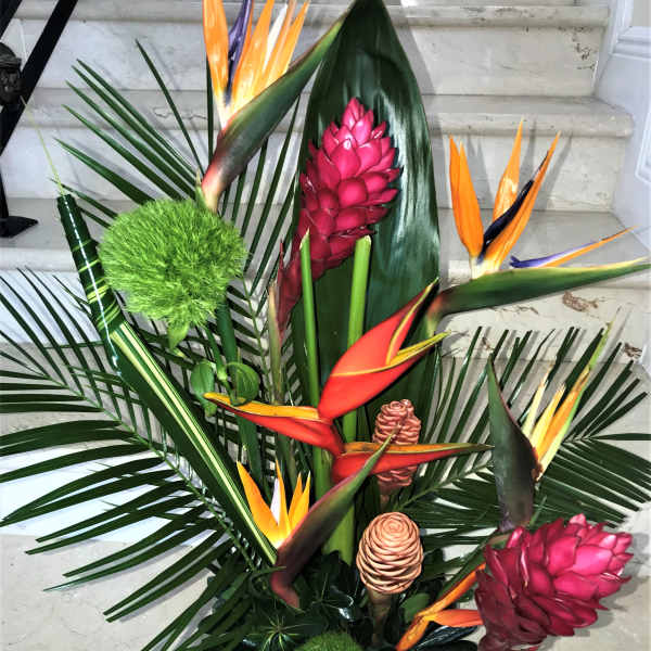 Tall tropical arrangement with orange blooms and pink gingers among large palm foliage in a black container