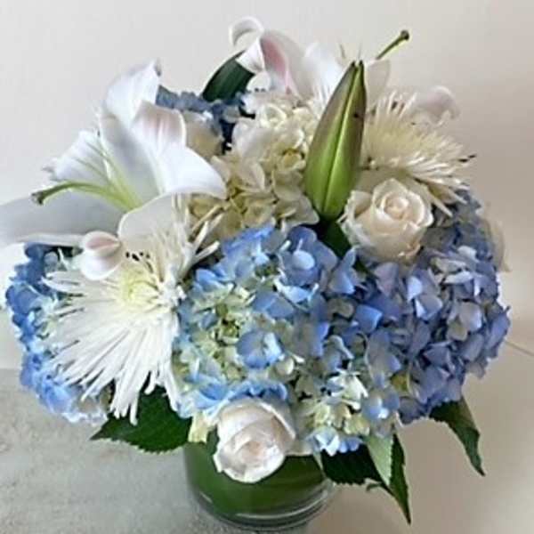 Round arrangement of blue hydrangeas, white lilies, roses, and mums in a clear glass vase