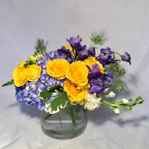Short glass vase of yellow roses, blue hydrangeas, and purple flowers in a compact mixed arrangement