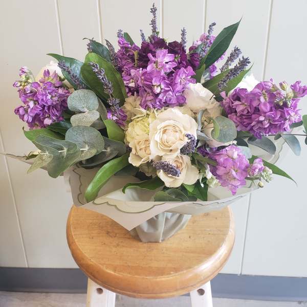Hand-tied bouquet of purple stock and cream roses wrapped in pale paper on a wooden stool.