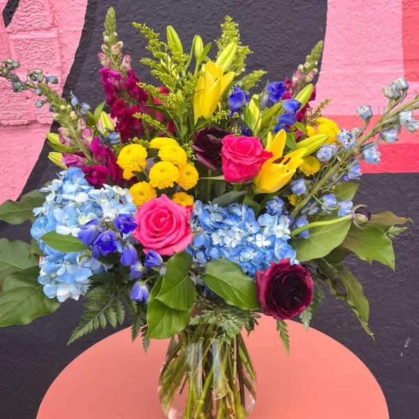 Colorful mixed bouquet with blue hydrangeas, yellow lilies, and pink roses in a clear glass vase.
