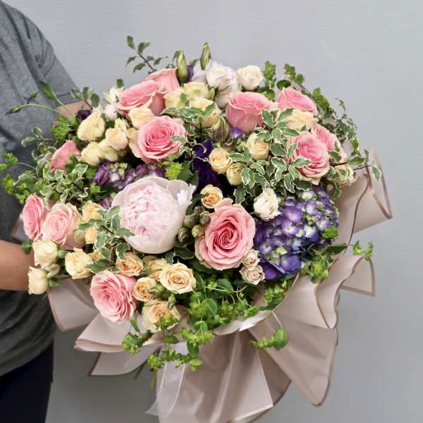 Large hand-tied bouquet of pink roses, cream flowers, and purple hydrangeas wrapped in blush paper