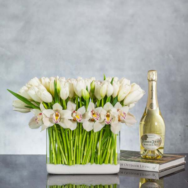 Rectangular vase of white tulips and orchids with a champagne bottle beside it on stacked books