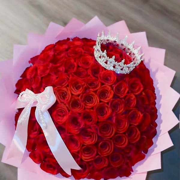Round bouquet of red roses wrapped in pink paper with a jeweled crown and white ribbon bow