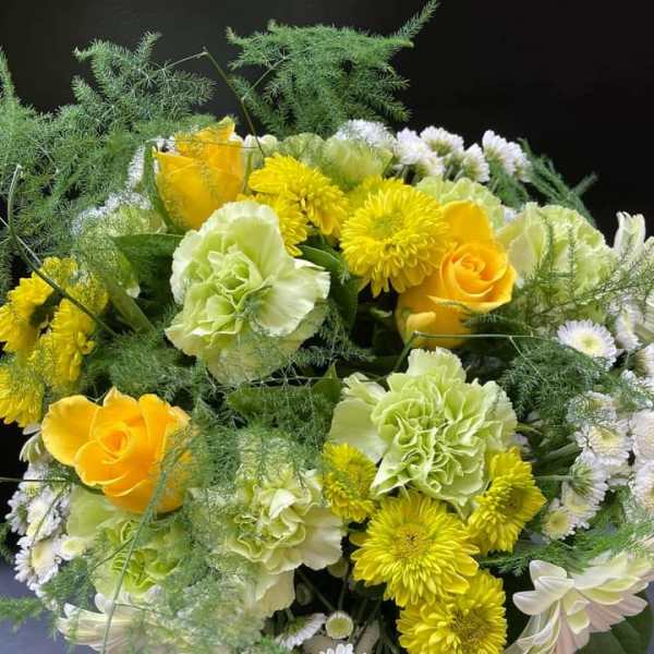 Round arrangement of yellow roses, green carnations, white chrysanthemums, and airy fern-like greenery.