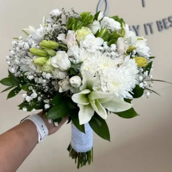 Hand-tied white bouquet with lilies, roses, and mums wrapped in white ribbon, held against a neutral background.