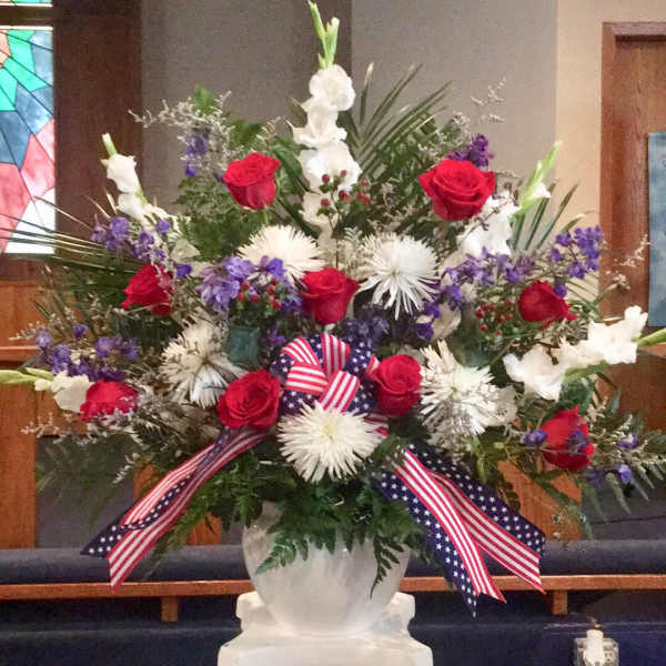 Tall red, white, and purple flower arrangement in a white urn with patriotic ribbon on a pedestal