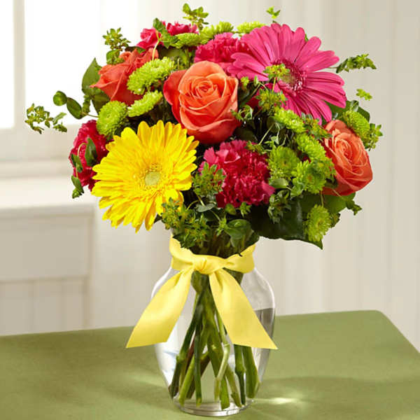 Mixed bouquet of roses, gerbera daisies, and carnations in a glass vase with a yellow ribbon