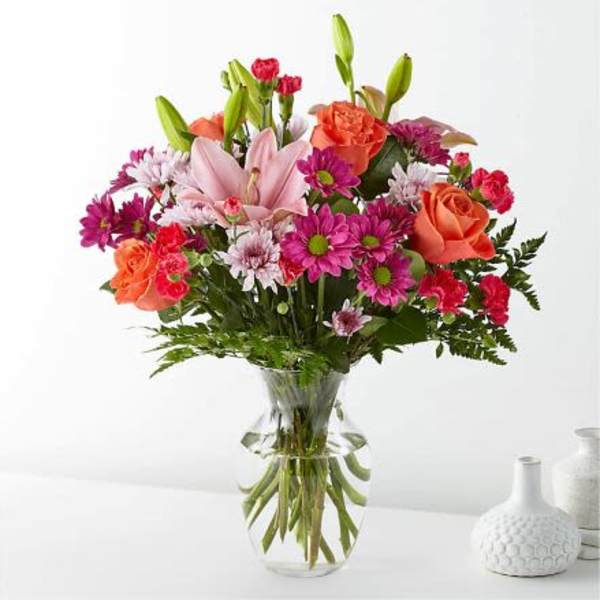 Mixed bouquet of pink lilies, orange roses, and bright daisies in a clear glass vase