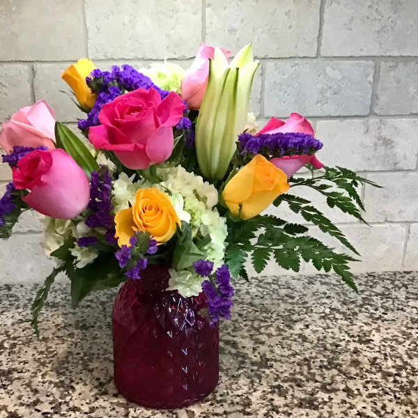 Mixed bouquet of pink and yellow roses, lilies and white blooms in a red glass vase on a speckled counter.
