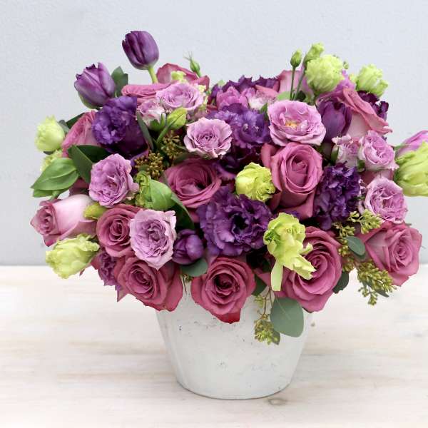 Round arrangement of mauve roses, purple lisianthus, and green blooms in a white pot