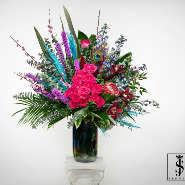 Tall arrangement of hot pink roses, orchids, protea, and peacock feathers in a dark glass vase