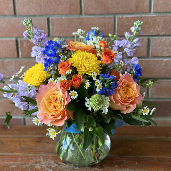 Mixed bouquet of orange roses, yellow mums, blue flowers and white daisies in a round glass vase