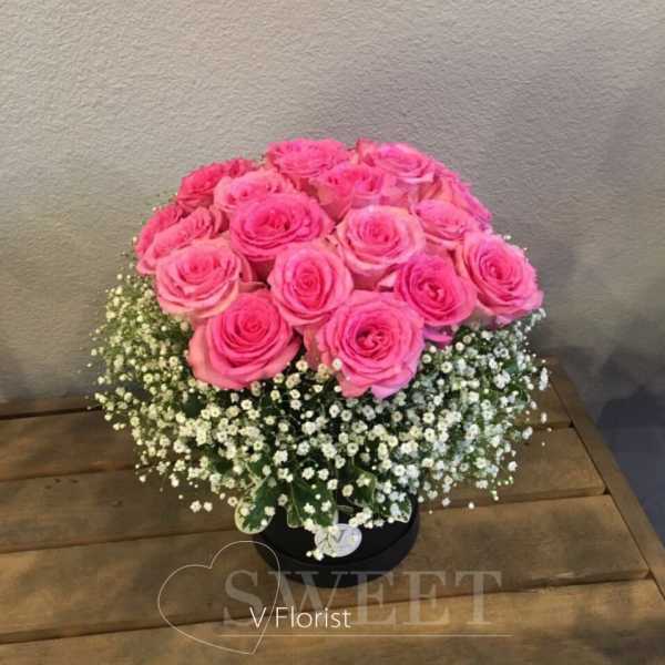 Round arrangement of bright pink roses with a collar of white filler flowers in a black hatbox.
