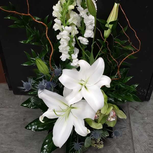 Tall arrangement of white lilies and snapdragons with blue thistle in a dark container
