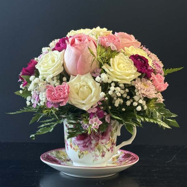 Compact arrangement of pink and white roses and mixed blooms in a floral teacup with matching saucer.