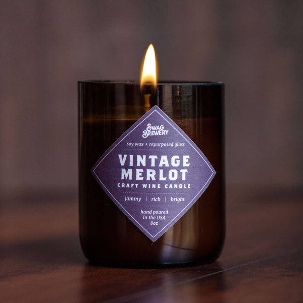 Brown glass jar candle labeled Vintage Merlot with a single flame on a wood surface