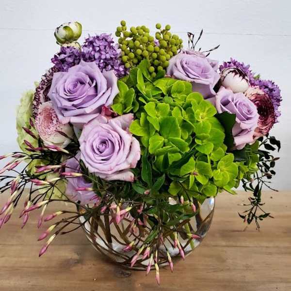 Round arrangement of lavender roses, green hydrangea, and mixed blooms in a clear glass bowl vase