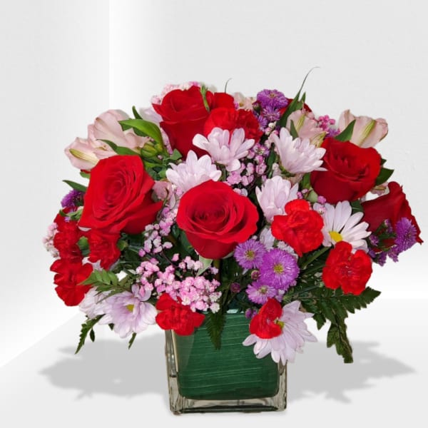 Mixed arrangement of red roses, red carnations and pastel blooms in a clear glass cube vase