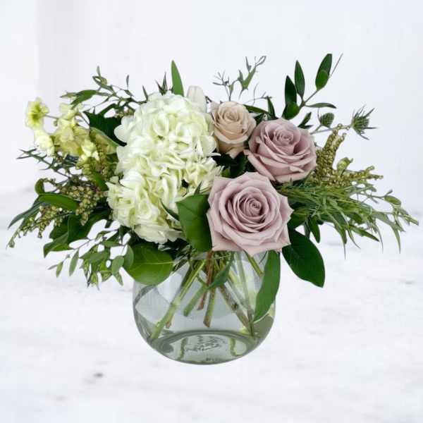 Low arrangement of white hydrangea and dusty lavender roses in a round glass vase