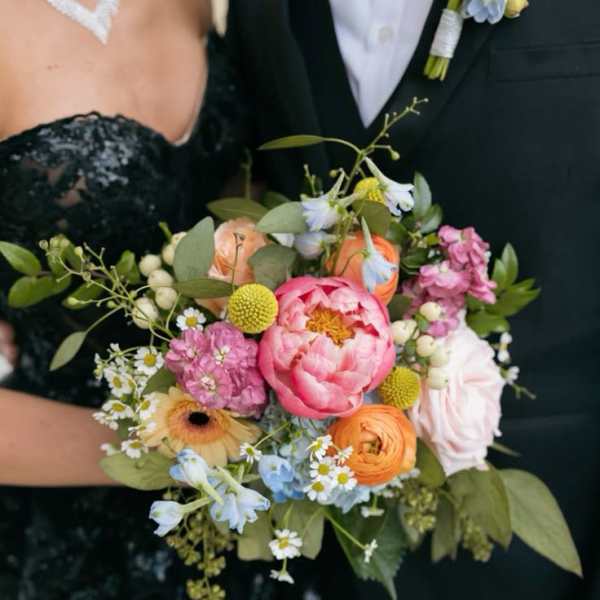 Colorful hand-tied bouquet of pink peonies, orange ranunculus, and mixed blooms held by a dressed-up couple