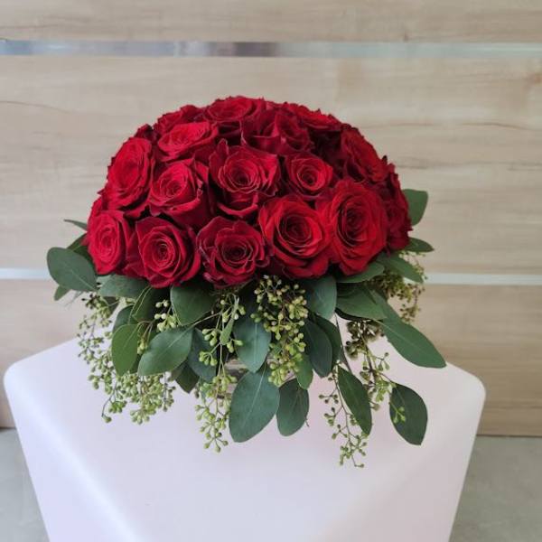 Low round arrangement of red roses with cascading greenery in a clear vase on a white pedestal