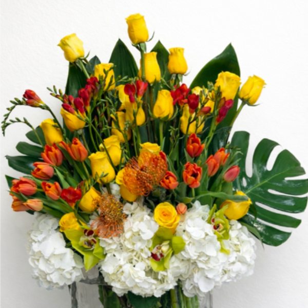 Tall arrangement of yellow roses, orange tulips, protea, orchids and white hydrangeas in a clear glass vase