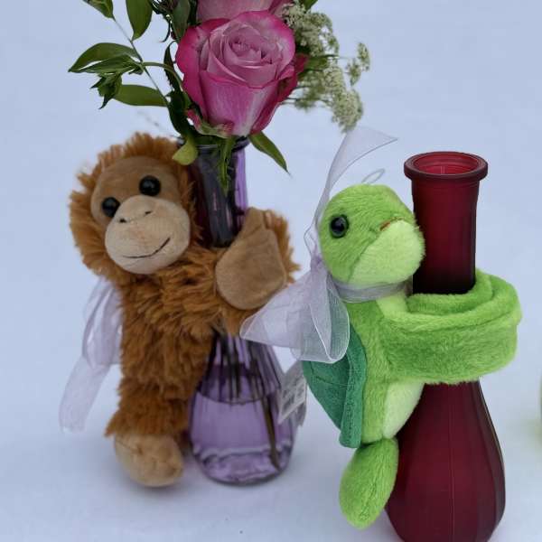 Pink roses in a purple bud vase with a monkey plush and a red vase hugged by a green turtle plush.