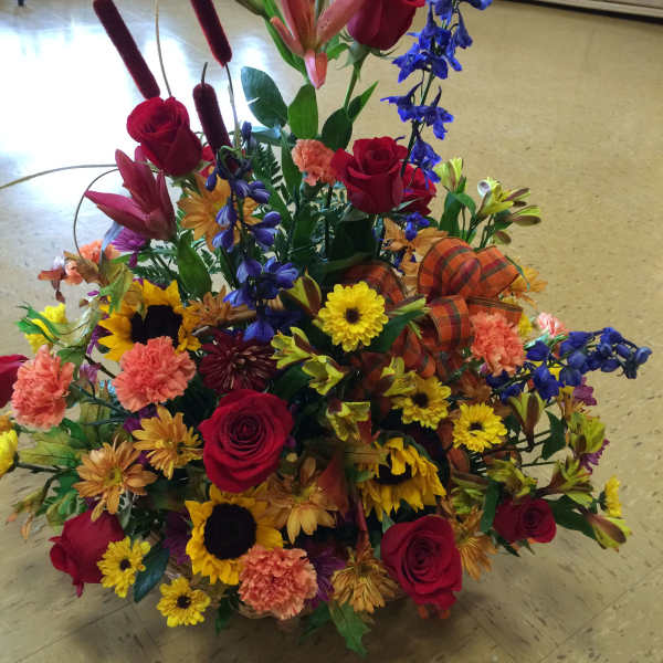 Tall mixed arrangement of red roses, sunflowers, lilies, and fall-colored blooms with a plaid bow.