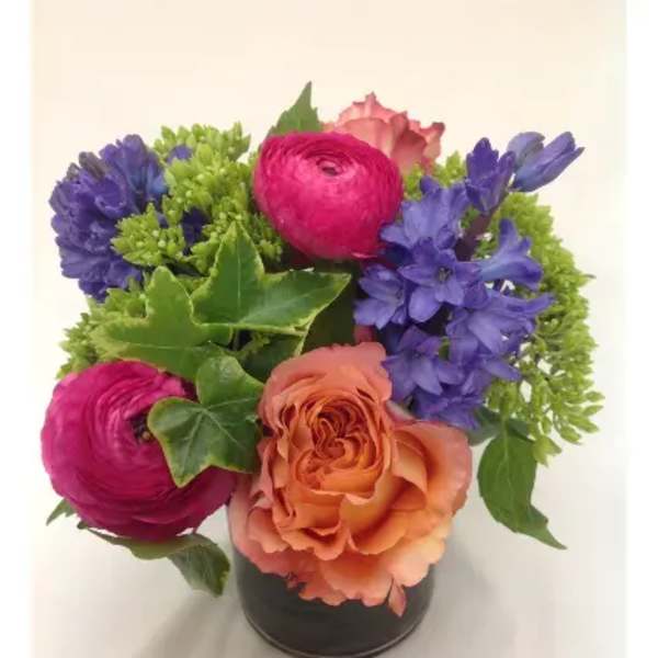 Short arrangement of peach rose, pink ranunculus, and purple flowers in a small glass vase