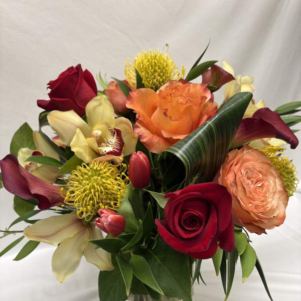 Vibrant arrangement of red and orange roses, yellow pincushion protea, orchids and tulips in a clear glass vase