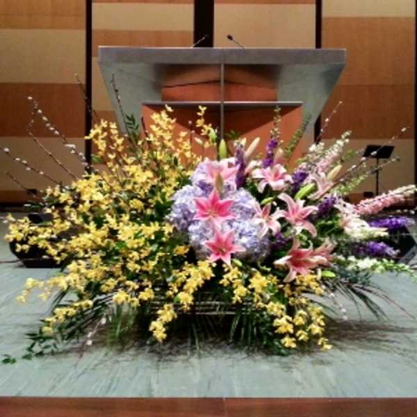 Large stage floral arrangement with pink lilies, lavender hydrangeas, yellow orchids, and tall accent stems