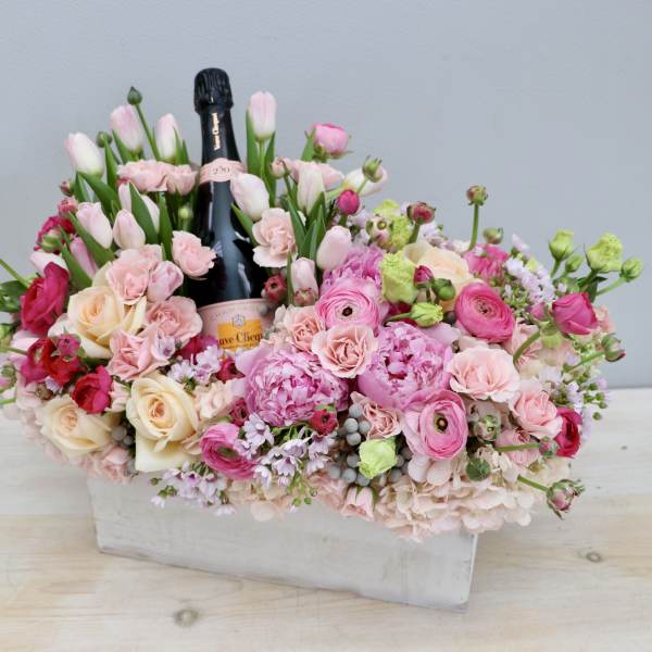 Pink and cream flower arrangement in a white box with a champagne bottle in the center