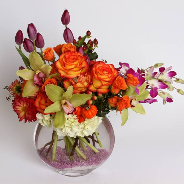 Mixed bouquet of orange roses, purple tulips, and orchids in a round glass vase